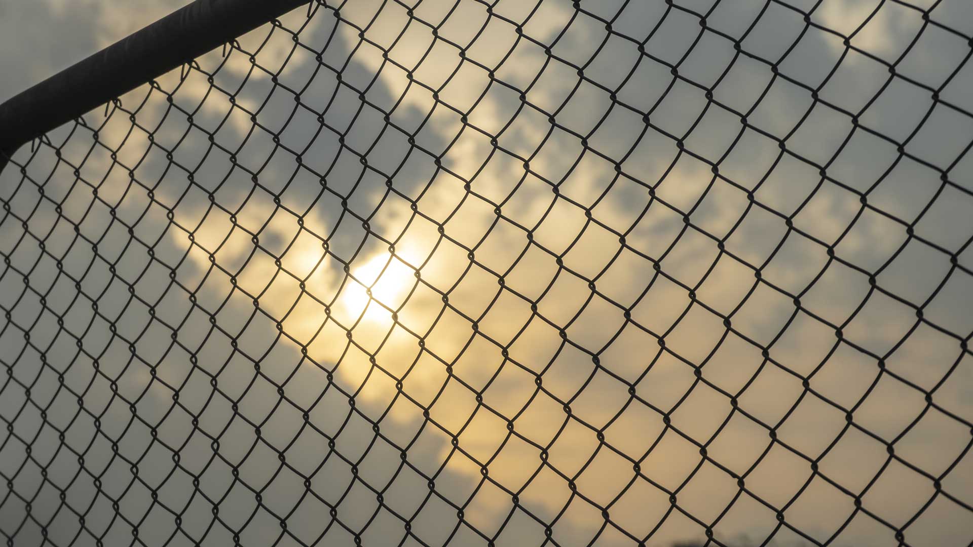 A black chain link fence is seen early in the morning with the sun hidden behind the clouds in the background