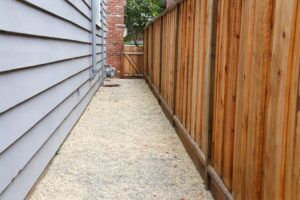 Long narrow side yard, new wood fence on the right with house on the left, home paint chipping and peeling. Meter by brick chimney exterior, fence to front yard with trees